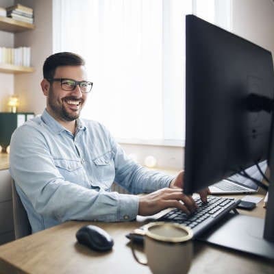 A white male with dark hair, a dark beard, and black framed glasses in a light blue button up shirt sits smiling at his desktop monitor. A black wireless mouse is beside him as well as a coffee mug.