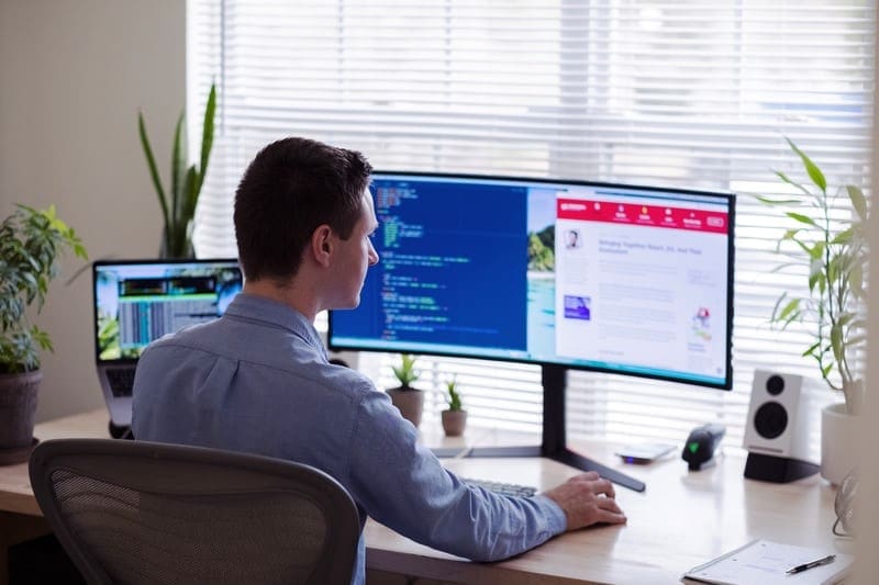 Male in a blut botton down shirt works on various monitors at a desk, surrounded by plants